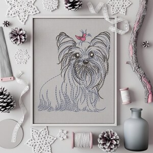 K&ouml;nnte beinhalten: Gerahmte Stickerei eines Yorkshire Terriers. Das Bild zeigt den Hund in Grau- und Blaut&ouml;nen, mit einem rosa Schmetterling. Dekorative Elemente wie Tannenzapfen und Spitze umgeben das Kunstwerk.