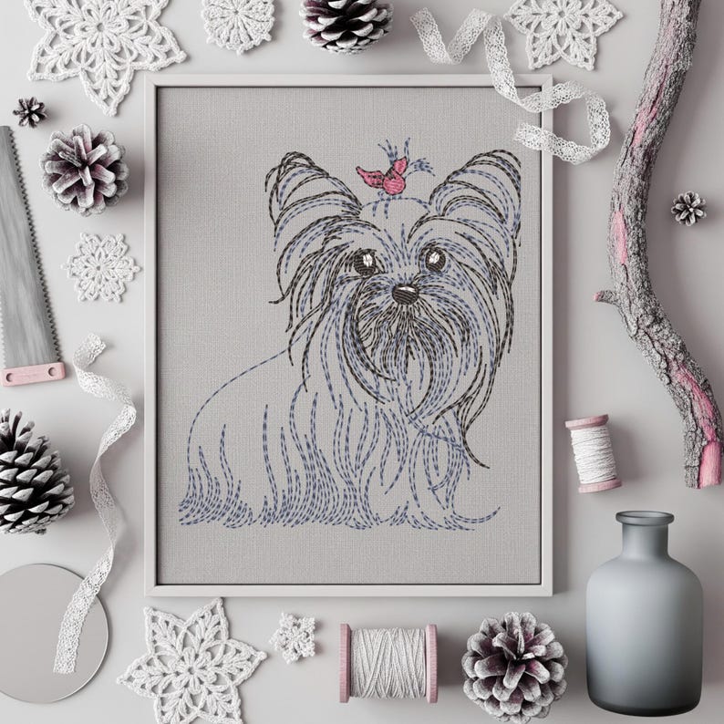 K&ouml;nnte beinhalten: Gerahmte Stickerei eines Yorkshire Terriers. Das Bild zeigt den Hund in Grau- und Blaut&ouml;nen, mit einem rosa Schmetterling. Dekorative Elemente wie Tannenzapfen und Spitze umgeben das Kunstwerk.
