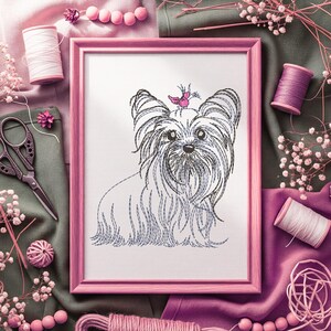 K&ouml;nnte beinhalten: Gerahmte Stickerei eines Yorkshire Terriers mit einem rosa Schmetterling auf dem Kopf. Der Hund ist in schwarzem und blauem Faden auf wei&szlig;em Hintergrund dargestellt, in einem rosa Rahmen. Das Bild ist von N&auml;hutensilien umgeben, darunter Garnrollen und Scheren.