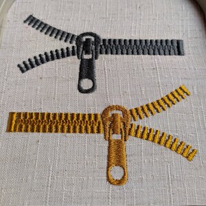 May include: Two embroidered zipper designs on a white fabric. One zipper is black and the other is gold.