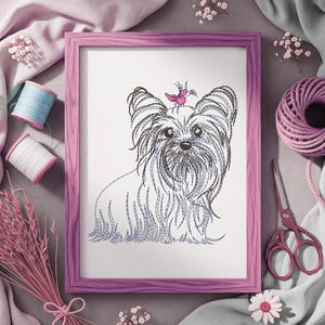 K&ouml;nnte beinhalten: Gerahmte Stickerei eines Yorkshire Terriers mit einem rosa Vogel auf dem Kopf. Der Hund ist in schwarzem und blauem Faden auf wei&szlig;em Hintergrund dargestellt, in einem rosa Holzrahmen. N&auml;hutensilien, darunter Garnrollen und Scheren, umgeben den Rahmen.