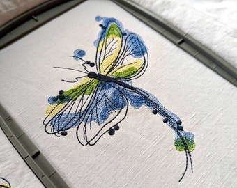 Op de afbeelding: Een borduurmotief met een blauwe en groene vlinder op witte stof in een borduurring.
