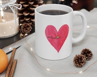 May include: White ceramic mug with a pink watercolor heart and the text "Love is love" with a rainbow heart. The mug is filled with coffee and steam is rising from the top.