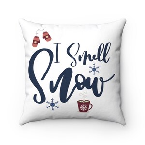 May include: White throw pillow with the text "I smell snow" in navy blue script. The pillow features a red mug with marshmallows and steam, two red mittens, and two snowflakes.