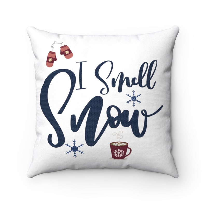 May include: White throw pillow with the text "I smell snow" in navy blue script. The pillow features a red mug with marshmallows and steam, two red mittens, and two snowflakes.