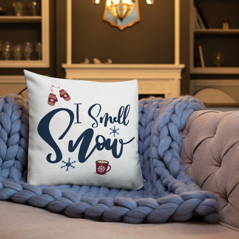 May include: White throw pillow with the text "I Smell Snow" in blue lettering. The pillow features a red and white mug with steam coming from it, two red mittens, and snowflakes.