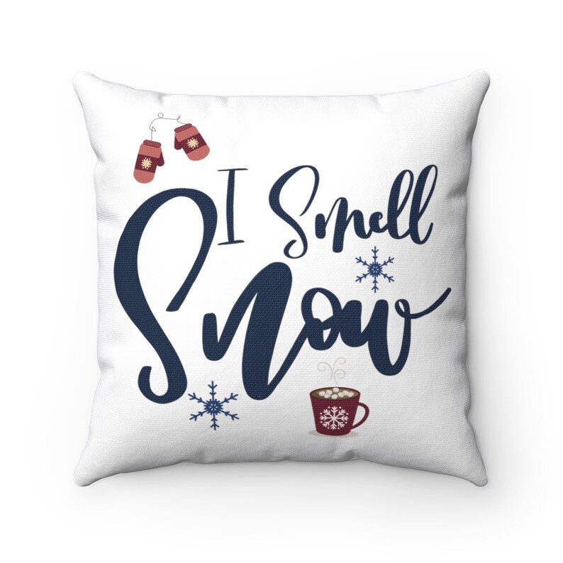 May include: White throw pillow with the text "I smell snow" in blue script. The pillow features a red mug with marshmallows and steam, two red mittens, and snowflakes.