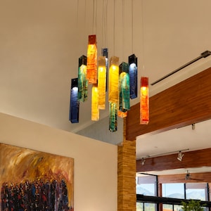 May include: A multi-colored glass pendant chandelier featuring rectangular, translucent hanging lights in shades of red, orange, yellow, blue, and green, suspended from the ceiling by thin wires above a wooden sideboard and a large abstract painting.