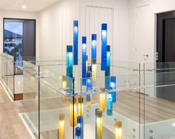 Könnte beinhalten: Ein Kronleuchter mit mehreren Anhängern, bestehend aus zahlreichen rechteckigen Glasstäben in Blau- und Bernsteintönen, die an dünnen Drähten von einer quadratischen Deckenhalterung hängen und eine vertikale Anordnung bilden, die den Raum beleuchtet.