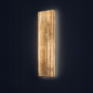 May include: A rectangular wall light fixture with a vertical orientation. The surface features a textured, mottled gold and brown finish that emits a soft, diffused glow from behind its edges, creating a halo effect against the dark background.