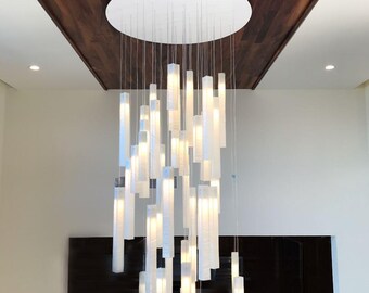 Op de afbeelding: Moderne kroonluchter met talrijke witte, rechthoekige lichtarmaturen die aan een cirkelvormige plafondbevestiging hangen. De lichten hangen op verschillende lengtes, wat een dynamisch visueel effect creëert. Het plafond is donker hout.