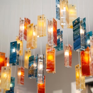 May include: A multi-pendant chandelier featuring numerous rectangular glass panels in shades of blue, orange, yellow, and red. Each glass piece is suspended by a thin wire and contains an internal light source, creating a warm, glowing effect.