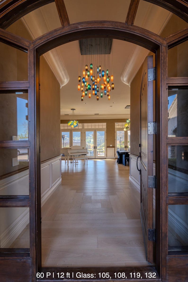 a spacious and well-lit entryway leading to a living room area. The wooden door is open, revealing a grand piano and a chandelier hanging from the ceiling. The room has large windows that allow natural light to flood in, creating a warm and inviting atmosphere.