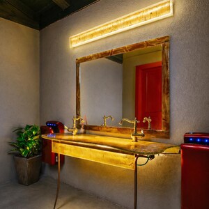 May include: A bathroom vanity with a large mirror, gold-toned fixtures, and a built-in light fixture. The vanity has a unique, organic shape and is supported by slender, curved legs. Red hand dryers are mounted on the wall.