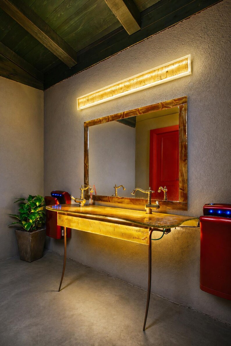May include: A bathroom vanity with a large mirror, gold-toned fixtures, and a built-in light fixture. The vanity has a unique, organic shape and is supported by slender, curved legs. Red hand dryers are mounted on the wall.