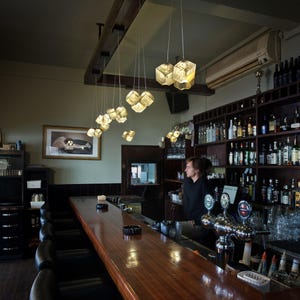 May include: A long wooden bar counter with black leather stools, illuminated by a series of hanging cube-shaped pendant lights. Behind the bar, shelves are stocked with various glass bottles, and a person stands near the beer tap system.