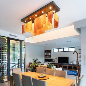 May include: A rectangular ceiling light fixture featuring multiple suspended, translucent amber and orange rectangular panels with a wood grain pattern, illuminated by integrated lights within a dark frame, hanging above a dining table.