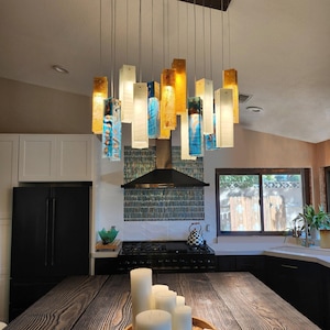 Pode incluir: Um lustre de múltiplos pendentes com blocos de vidro retangulares em tons de âmbar, azul e branco, suspensos por um suporte de teto retangular escuro com fios finos, criando uma luminária em camadas com alturas variadas.