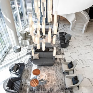 May include: A high-angle view of a lobby featuring a large, multi-tiered vertical light fixture with rectangular glass shades. The space includes gray sofas, dark gray and light gray armchairs, round wooden side tables, and a brown tufted bench.
