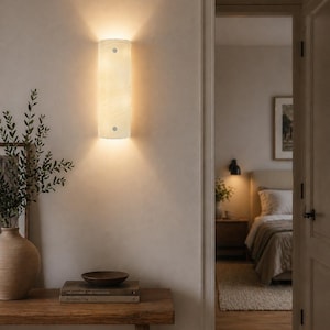 May include: A rectangular, cream-colored wall sconce with a soft glow, mounted on a light-colored wall. The sconce is positioned above a wooden table with a vase, books, and a bowl. The room is softly lit, with a glimpse of a bedroom in the background.