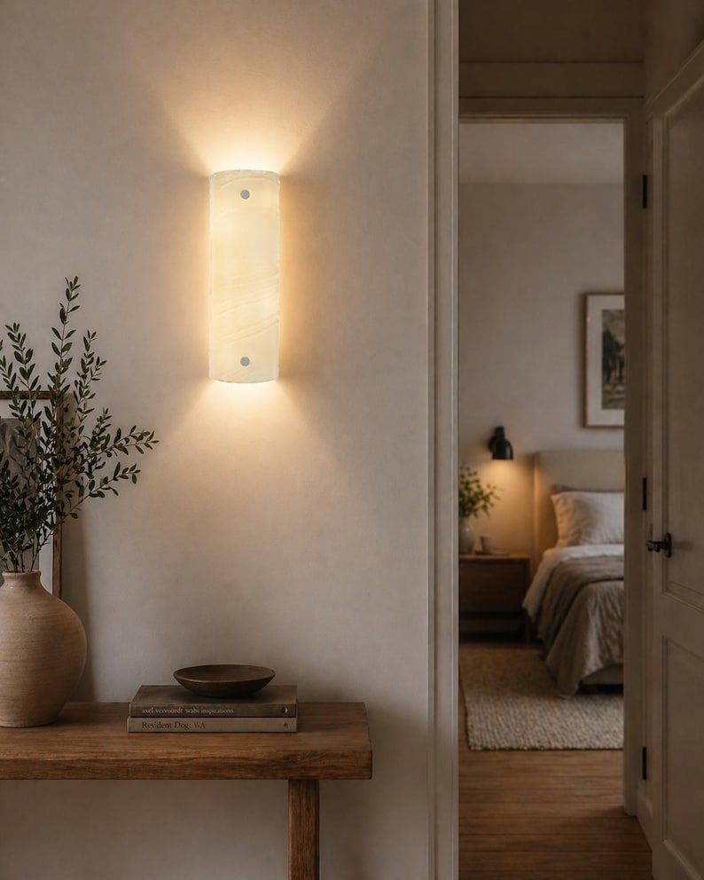 May include: A rectangular, cream-colored wall sconce with a soft glow, mounted on a light-colored wall. The sconce is positioned above a wooden table with a vase, books, and a bowl. The room is softly lit, with a glimpse of a bedroom in the background.