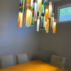 a modern dining room with a wooden table and six chairs. The table is illuminated by a hanging chandelier composed of colorful glass rods that cast a warm glow throughout the room.