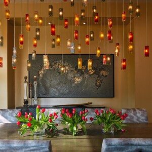 a modern dining room with a wooden table, gray upholstered chairs, and a large wall art piece. The table is set with red tulips in vases, and the room is illuminated by hanging lights.