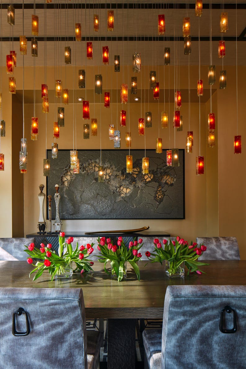 a modern dining room with a wooden table, gray upholstered chairs, and a large wall art piece. The table is set with red tulips in vases, and the room is illuminated by hanging lights.