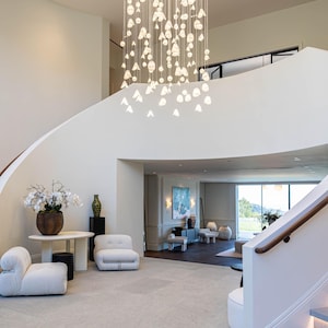 a spacious and modern living room with a large, elegant chandelier hanging from the ceiling, creating a warm and inviting atmosphere. The room is furnished with white sofas and chairs, as well as a dining table, all arranged to create a comfortable and functional space. The overall design and layout of the room suggest a high-end, luxurious living environment.