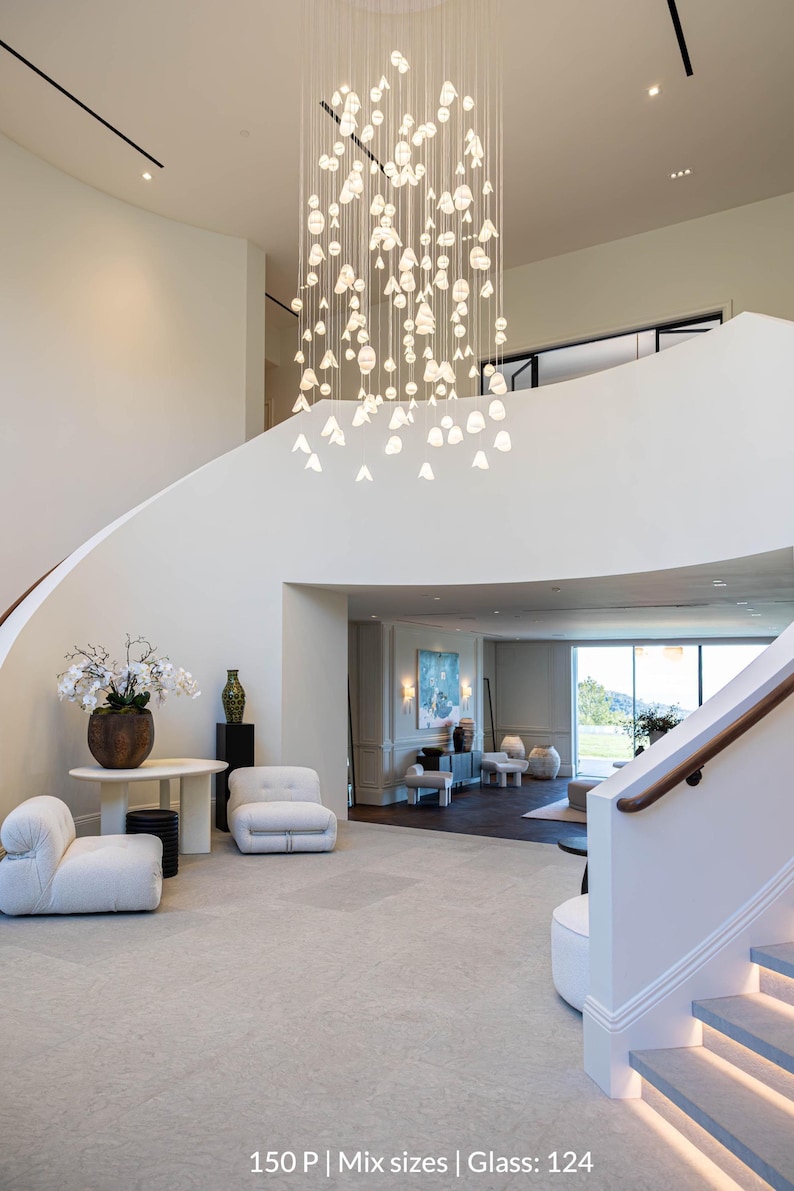 a spacious and modern living room with a large, elegant chandelier hanging from the ceiling, creating a warm and inviting atmosphere. The room is furnished with white sofas and chairs, as well as a dining table, all arranged to create a comfortable and functional space. The overall design and layout of the room suggest a high-end, luxurious living environment.