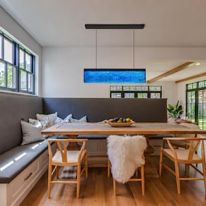 Puede incluir: Una lámpara colgante rectangular con un panel de vidrio azul vibrante suspendido por cables negros delgados sobre una mesa de comedor de madera. La luz presenta un marco negro elegante y emite un brillo azul intenso en la habitación.