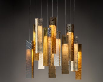 Op de afbeelding: Een moderne kroonluchter met rechthoekige, verticale armaturen in verschillende tinten goud, zilver en wit. De armaturen hangen aan dunne draden, waardoor een gelaagde, artistieke weergave ontstaat. Het algehele ontwerp is eigentijds en elegant.