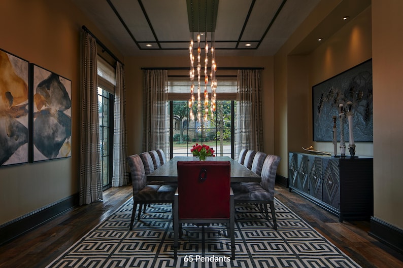 a luxurious dining room with a large wooden table surrounded by gray chairs, a red chair, and a chandelier with multiple lights hanging above the table. The room features a large window with curtains, and there are paintings on the walls. The overall design and decor create an elegant and sophisticated atmosphere.