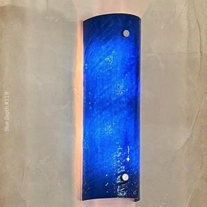 May include: A wall sconce with a curved, cylindrical design. The sconce is a deep blue color with a textured appearance, and is mounted with two silver-colored fasteners. The text "Blue Depth #119" is visible.