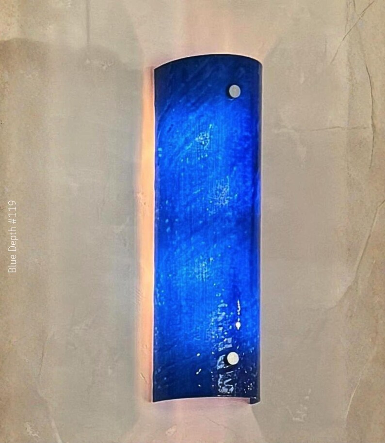 May include: A wall sconce with a curved, cylindrical design. The sconce is a deep blue color with a textured appearance, and is mounted with two silver-colored fasteners. The text "Blue Depth #119" is visible.