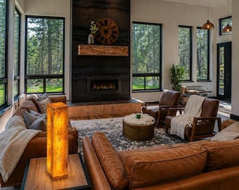 May include: A tall, rectangular, glowing amber-colored lamp sits on a square wooden side table with a black metal frame. The lamp emits a warm, soft light, illuminating the surrounding living room area with brown leather sofas and chairs.