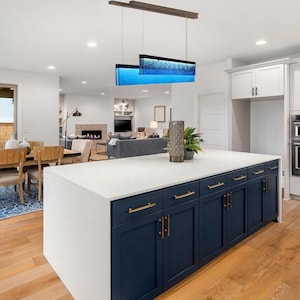 May include: A kitchen island with a white countertop and dark blue cabinets featuring gold hardware. Above the island hangs a rectangular pendant light with a blue glass panel. A tall, textured, cylindrical vase sits on the white surface.
