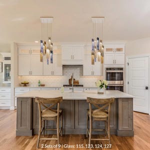a modern kitchen with a large island, two wooden chairs, and a sink. The kitchen has white cabinets and a marble countertop. The space is illuminated by two hanging pendant lights, and there is a vase of flowers on the countertop.