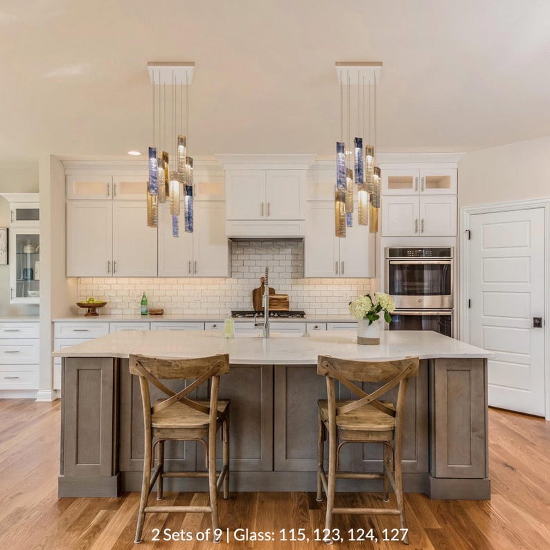 a modern kitchen with a large island, two wooden chairs, and a sink. The kitchen has white cabinets and a marble countertop. The space is illuminated by two hanging pendant lights, and there is a vase of flowers on the countertop.