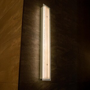 May include: A long, rectangular wall sconce with a bright, warm light. The fixture is encased in a clear, rectangular frame and mounted on a dark, textured wall. The sconce is a vertical design, providing a modern lighting element.
