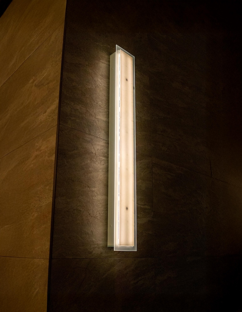 May include: A long, rectangular wall sconce with a bright, warm light. The fixture is encased in a clear, rectangular frame and mounted on a dark, textured wall. The sconce is a vertical design, providing a modern lighting element.