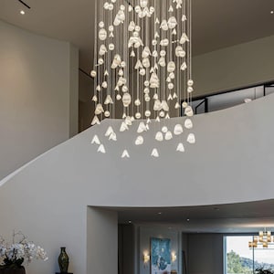 May include: A modern chandelier with numerous white, organic-shaped light fixtures suspended from the ceiling. The lights are arranged in a cascading pattern, illuminating a bright, open space with a curved staircase and minimalist decor.