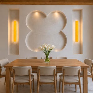 May include: A dining room with a wooden table and six beige chairs. The room features a white wall with a clover-shaped design and two vertical yellow lights. A vase of white flowers sits on the table, creating a bright and inviting space.