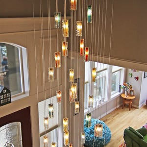 a spacious living room with a high ceiling, where a large chandelier with numerous hanging lights is suspended. The room is furnished with a green armchair, a wooden coffee table, and a rug on the floor. The overall atmosphere of the room is warm and inviting, with the chandelier providing a striking focal point.