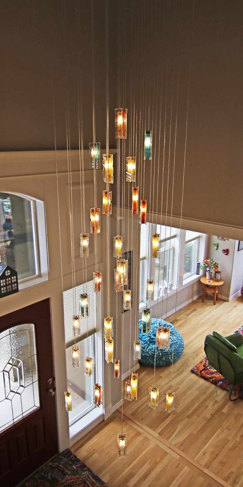 a spacious living room with a high ceiling, where a large chandelier with numerous hanging lights is suspended. The room is furnished with a green armchair, a wooden coffee table, and a rug on the floor. The overall atmosphere of the room is warm and inviting, with the chandelier providing a striking focal point.