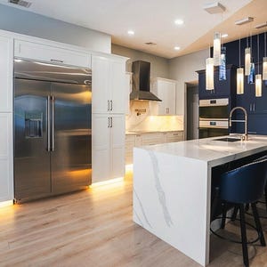 a modern, spacious kitchen with a large central island, white cabinets, and stainless steel appliances. The kitchen features a marble countertop and a sink, as well as a refrigerator and an oven. The space is illuminated by pendant lights hanging above the island, creating a warm and inviting atmosphere.