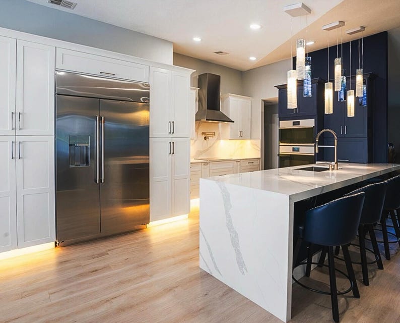 a modern, spacious kitchen with a large central island, white cabinets, and stainless steel appliances. The kitchen features a marble countertop and a sink, as well as a refrigerator and an oven. The space is illuminated by pendant lights hanging above the island, creating a warm and inviting atmosphere.