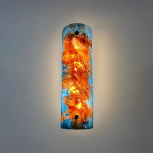 Puede incluir: Aplique de pared rectangular con un diseño abstracto azul y naranja. El aplique tiene una parte superior e inferior redondeadas con dos agujeros. El interior brilla con luz cálida, creando una lámpara decorativa.