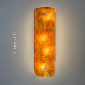 May include: A cylindrical wall sconce with a honey-colored, textured surface. The sconce has a warm, glowing interior with three visible light sources. The text "Honey #101" is printed vertically on the left side of the image.