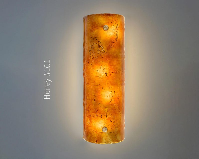 May include: A cylindrical wall sconce with a honey-colored, textured surface. The sconce has a warm, glowing interior with three visible light sources. The text "Honey #101" is printed vertically on the left side of the image.
